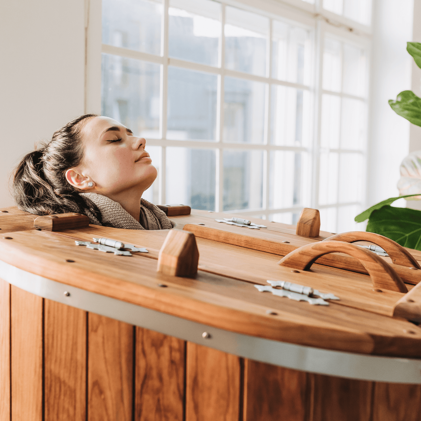 Vrouw zit in een houten stoomsauna met haar hoofd buiten de sauna en ontspant in een lichte ruimte bij het raam
